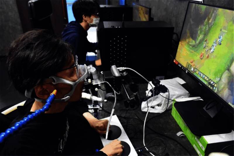 一名行动不便的老年人使用口含式操纵杆操控游戏角色,身旁助手协助调试设备,桌上摆放3D打印的定制手柄