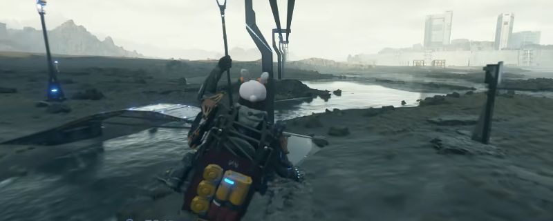 《死亡搁浅2》游戏中玩家手持改装后的消音狙击枪，在荒原环境中准备瞄准远处目标，枪械细节清晰可见